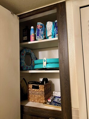 A storage cabinet with shelves containing various items including a blue inflatable ring, a wicker basket, and several toiletry bags. - Condo • 2 Beds •1 ba near Downtown-Riverwalk, La cantera-Fiesta Texas, Sea World (San Antonio)