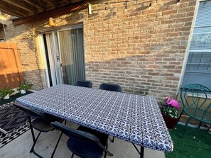 Um pátio com mesa, cadeiras, um muro de tijolos, uma porta de madeira e uma cadeira verde.