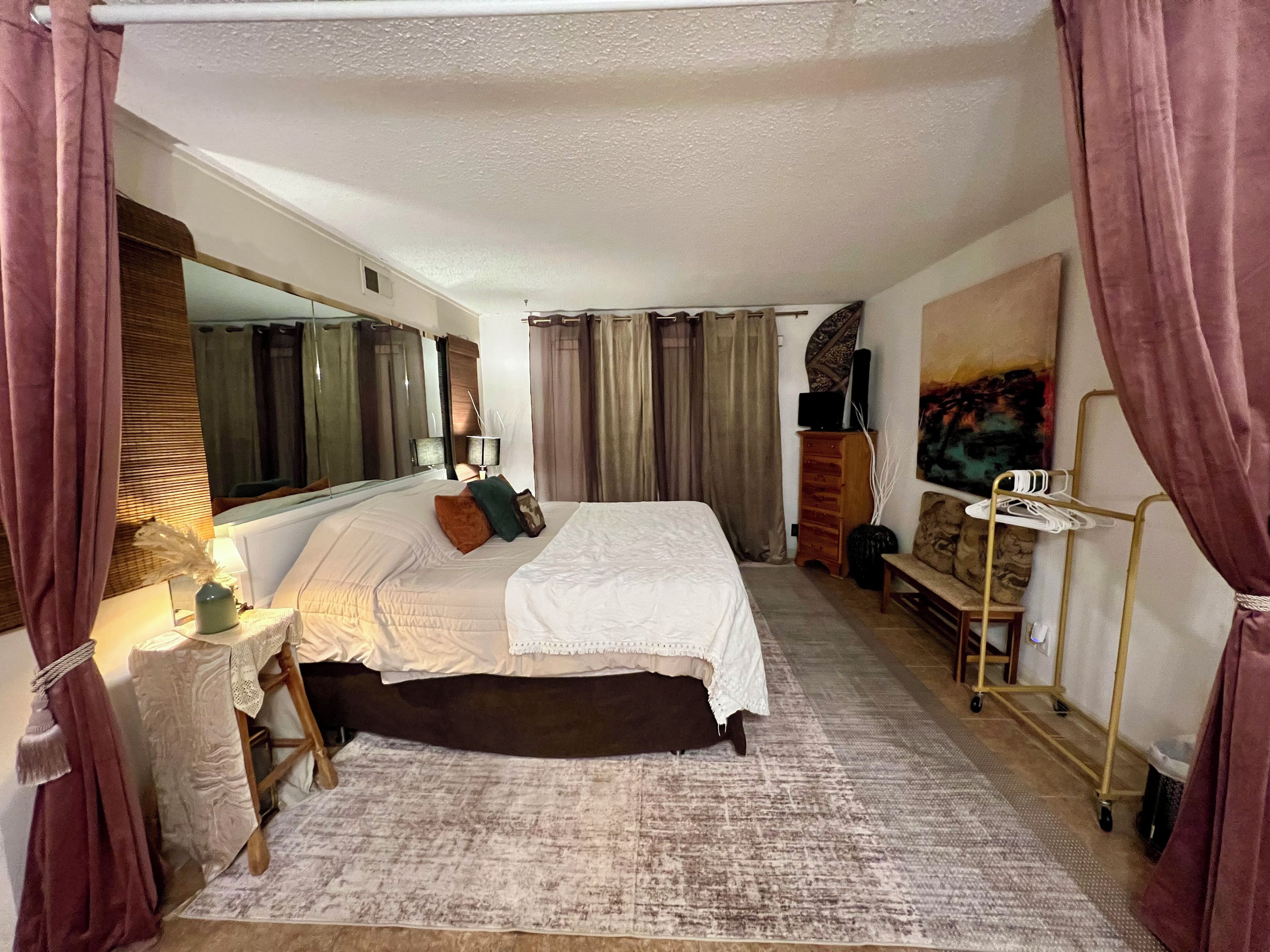Ein Hotelzimmer mit einem großen Bett, einem Nachttisch mit Lampe, einem Schminktisch, einem Sessel, einem kleinen Tisch ebenfalls mit Lampe, einem Spiegel, einem Bild an der Wand und einem Fenster mit Vorhängen.