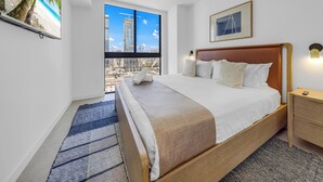 Ein Hotelzimmer mit einem großen Bett, Blick auf die Stadt, einem Gemälde an der Wand und einem Fernseher, der eine Strandansicht zeigt.