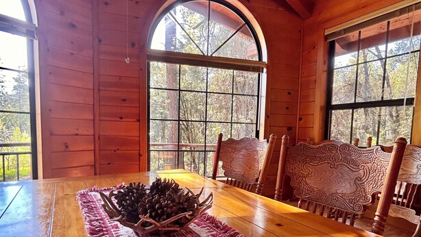 Interior de una cabaña de madera con una mesa de comedor, sillas, un ventilador de techo y ventanas amplias.
