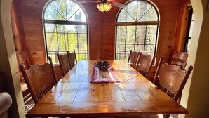 Una mesa de comedor de madera con sillas, un ventilador de techo y ventanas amplias con vistas a árboles.