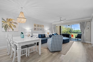 A modern living room with a white dining table, blue sofa, patterned ottoman, and a ceiling fan.