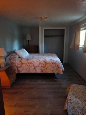 Une chambre à coucher comprenant un lit, une table de chevet, une commode, une lampe, un placard et une fenêtre.