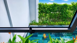 Piscina interior moderna com paredes de vidro transparente, rodeada por plantas tropicais exuberantes e vista para um denso conjunto de sebe verde lá fora.