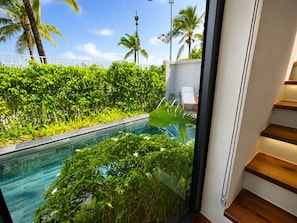 A modern indoor pool with glass walls, surrounded by lush greenery and palm trees, leading to a wooden staircase. - Kalia Villa (Phu Quoc)