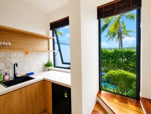 A kitchen with a black dishwasher, wooden cabinets, a white countertop, and a window with a view of a pool and palm trees. - Kalia Villa (Phu Quoc)