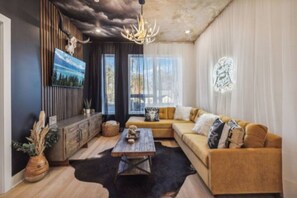Ein Wohnzimmer mit einem Ecksofa, einem Holztisch, einem aufgehängten Hirschgeweih und einem großen Fenster.
