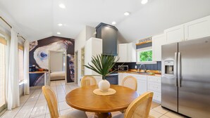 A modern kitchen with white cabinets, a stainless steel refrigerator, and a dining area with a wooden table and chairs. - Nautigull- A Fisherman's Delight (Apalachicola)