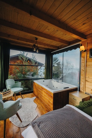 A room with a wooden ceiling, a large window, a bathtub, a chair, a bed, and a small table.