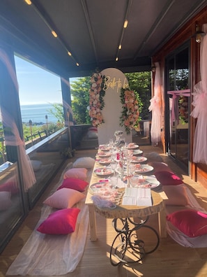 A dining area with a view of the ocean, a floral arch, and a table set for a meal.