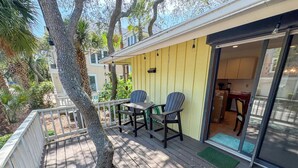 Una terraza de madera con dos sillas y una mesa, un edificio amarillo con puerta corredera de vidrio, y un árbol en primer plano.