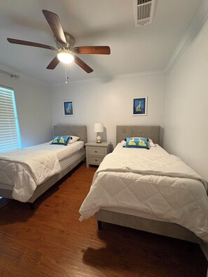 A bedroom with two single beds, a ceiling fan, a nightstand with a lamp, and two wall-mounted pictures. - Peaceful 4-bedroom house with AC in Baton Rouge . Bring your whole family (Baton Rouge)