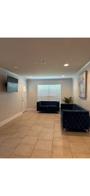 A spacious room with beige tiled flooring, a dark blue sofa, a television mounted on the wall, and a window with blinds. - Peaceful 4-bedroom house with AC in Baton Rouge . Bring your whole family (Baton Rouge)