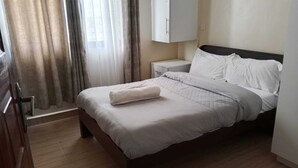 Ein ordentlich bezogenes Bett mit weißer Daunendecke und gefaltetem Handtuch, ein Nachttisch, ein Kleiderschrank und ein Fenster mit Vorhängen.
