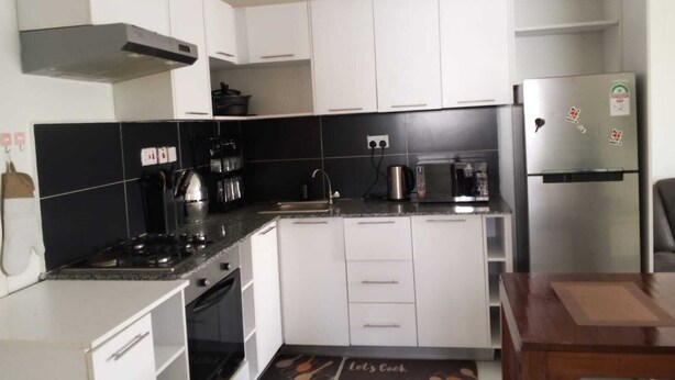 Eine kompakte Küche mit weißen Schränken, einer schwarzen Fliesenspiegel und einem Edelstahl-Kühlschrank sowie einer Kaffeemaschine.