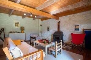 A cozy living room with a stone wall, wooden beams, a fireplace, a sofa, a table, and a red chair.