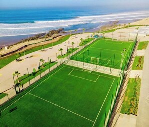 Eine Vogelperspektive auf einen Sportkomplex mit einem grünen Spielfeld, einem Tor und einem Strand im Hintergrund.