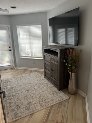 Ein Zimmer mit einem an der Wand befestigten Flachbildfernseher, einem Holzschrank, einem gemusterten Teppich und einer Vase mit Grünpflanzen.