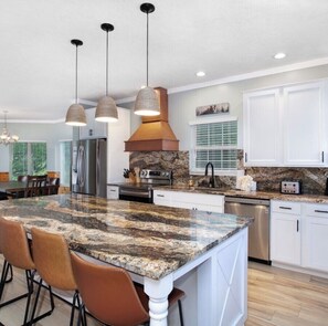 A kitchen with granite countertops, white cabinetry, a central island with bar stools, stainless steel appliances, pendant lighting, and a stone backsplash.