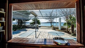A view through a wooden-framed window of a patio area with a metal pergola, stone tiles, and a metal railing overlooking a body of water. - Sea Side Moretti Near Vodice & Beach (Vodice)