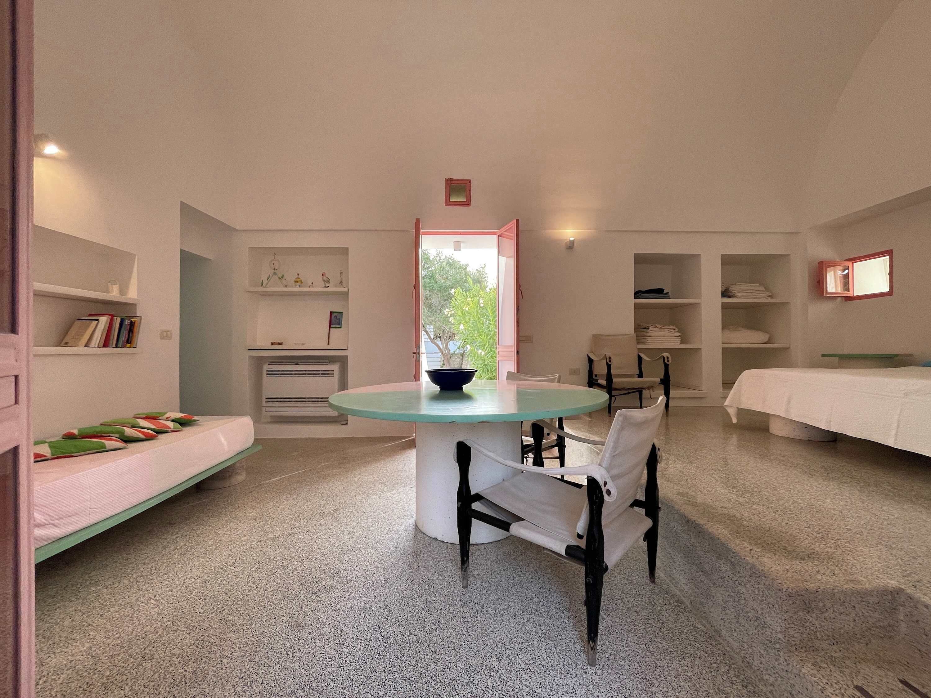 A room with a round table, chairs, a bed, a shelf with books, and a window.