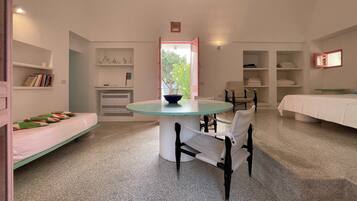 A room with a round table, chairs, a bed, a shelf with books, and a window.