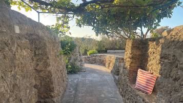 Un étroit sentier de pierre, bordé de murs de pierre bruts, mène à un espace ombragé où se trouve un coussin à rayures.