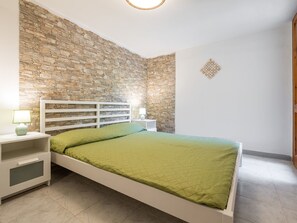 Une chambre avec un mur en pierre, un lit blanc, une table de chevet avec une lampe et un petit objet décoratif accroché au mur.