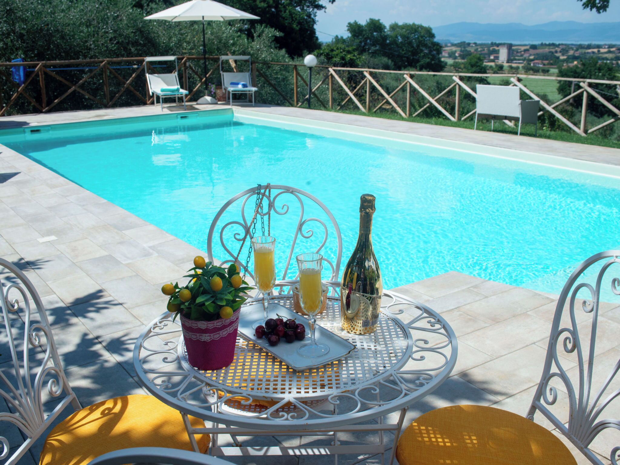 Une table au bord de la piscine avec des boissons, une bouteille et des fruits.