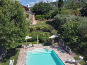 En swimmingpool med en terrasse, omgivet af frodigt grønt og en bygning i sten i baggrunden.