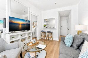 Un salon moderne avec un téléviseur, une table basse en verre, un canapé et un coin cuisine avec des tabourets.