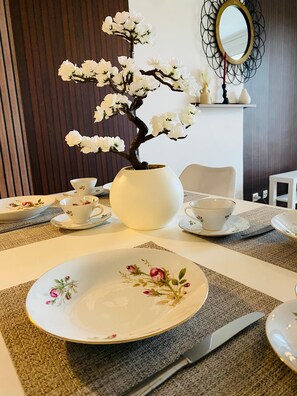 Una mesa de comedor con un centro de mesa floral, tazas de té blancas, platillos y un jarrón blanco.