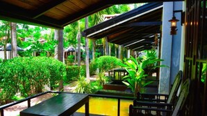 Um espaço ao ar livre coberto, com uma mesa e cadeiras, rodeado por vegetação exuberante e um pequeno elemento aquático.
