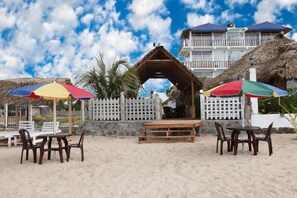 Un espacio al aire libre frente a la playa con mesas y sillas de madera, una estructura con techo de paja y una cerca blanca.
