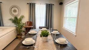 A dining room with a dark wooden table set for four, a clock on the wall, and a window with blinds.