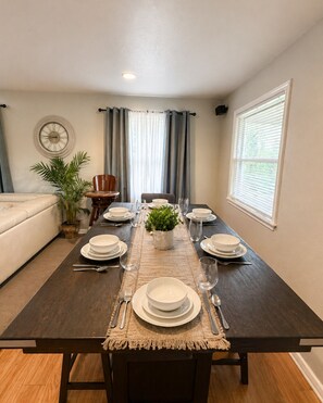 A dining room with a dark wooden table set for four, a clock on the wall, and a window with blinds.