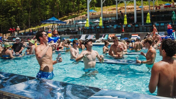 A swimming pool with people in the water, a covered area with umbrellas, and trees in the background.