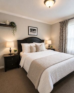 Une chambre à coucher bien rangée, avec un lit, deux tables de chevet dotées de lampes, un tableau accroché au mur et une plante suspendue.