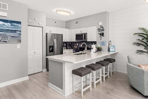 A modern kitchen with white cabinetry, a central island with bar stools, a stainless steel refrigerator, and a microwave. - Shore of Panama Unit 924,  BEACHFRONT 2 bd+Bunk, 3 bath, Sleeps 8 (Panama City Beach)