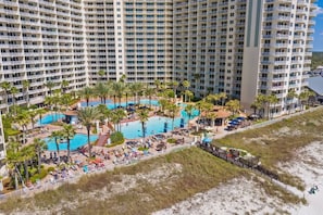 A high-rise hotel with multiple balconies, a central swimming pool surrounded by palm trees, and a beach area. - Shore of Panama Unit 924,  BEACHFRONT 2 bd+Bunk, 3 bath, Sleeps 8 (Panama City Beach)