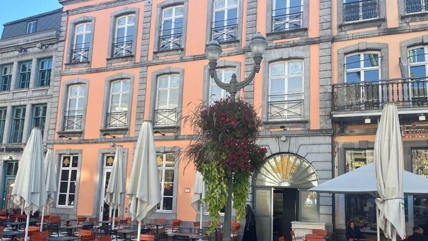 An outdoor café with tables and chairs, a street lamp, and a building with multiple windows and balconies. - HÔTEL GRAND PLACE (Mons)