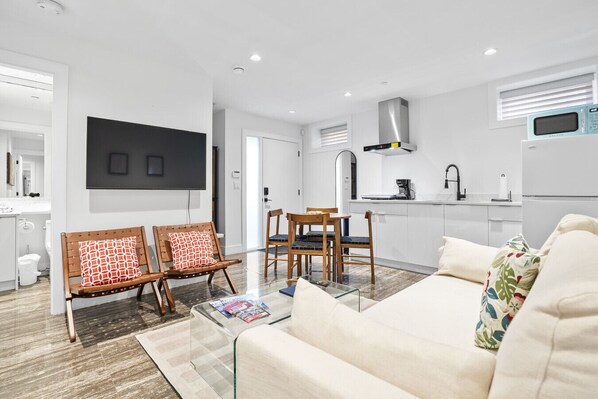 Una sala de estar moderna con sofá, una mesa de centro y televisor. Se observa un comedor con sillas y una mesa. La cocina es visible, con un microondas y una estufa.