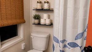 Un baño con un inodoro, dos estantes con artículos de aseo, una ventana con persianas y una cortina de ducha con motivos de peces.