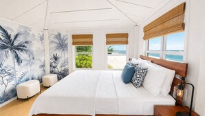 Un dormitorio con cama, dos ventanas que dan a la playa, un mural en la pared y una mesita de noche con lámpara.