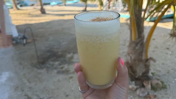 Una mano sosteniendo un vaso con una bebida, palmeras y una playa al fondo.