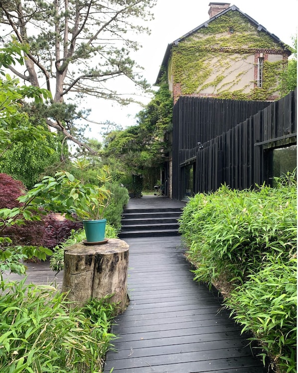 Un chemin en bois mène à une maison entourée d’une clôture noire et bordée de verdure de chaque côté.