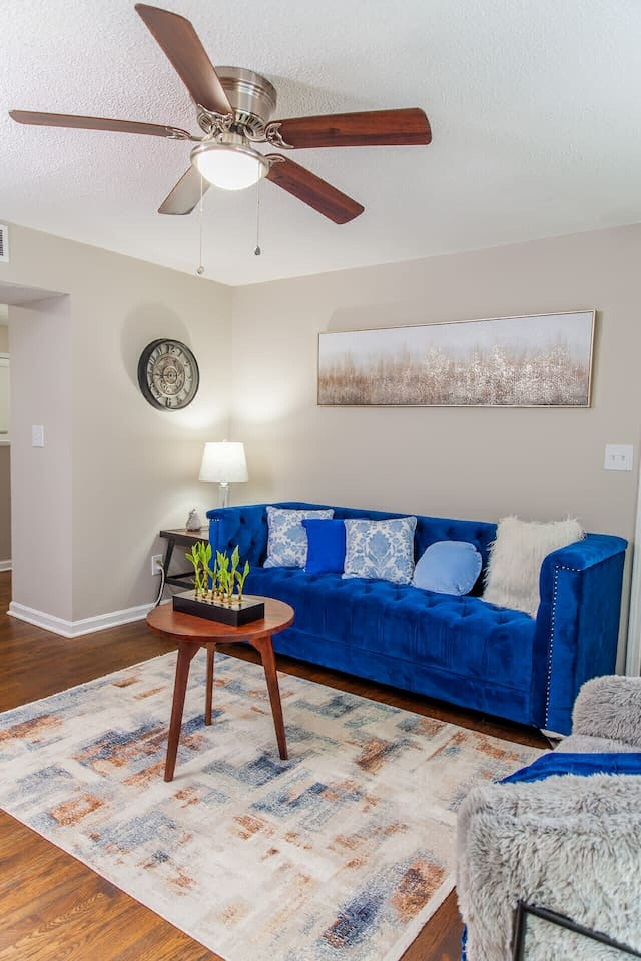 Un salon avec un canapé bleu, une table basse en bois, un ventilateur de plafond, une horloge murale et un tableau.