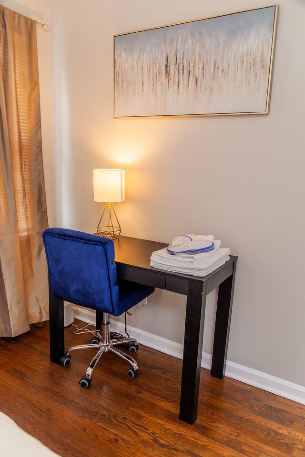 A room with a desk, chair, lamp, and a painting on the wall.