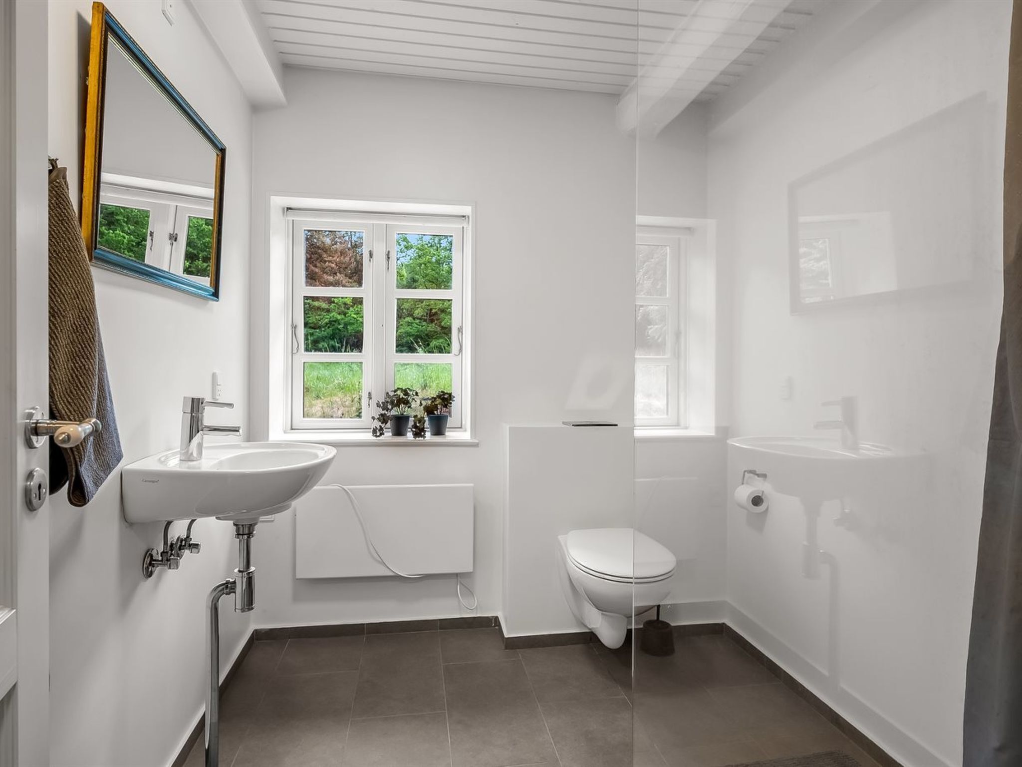 A bathroom with a white sink, a toilet, a window with a view of greenery, and a mirror with a blue frame.